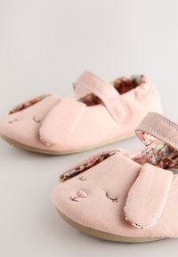 Vaaleanpunaiset kangastossut, joissa on pupun piirteitä, kuten kirjotut silmät ja nenä, pehmeät korvat ja kukkakuvioinen vuori; ne suljetaan tarranauhalla.