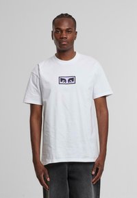 Hvid bomulds t-shirt med et centralt sort og lilla grafisk design. Korte ærmer, rund hals, afslappet pasform, ingen synlige mønstre.