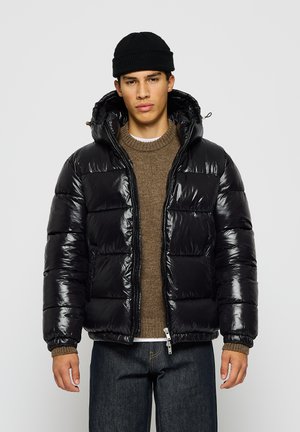 Giacca a vento nera in piuma d'oca con una texture lucida e trapuntata, cappuccio e polsini elasticizzati, indossata sopra un maglione marrone e abbinata a jeans scuri.