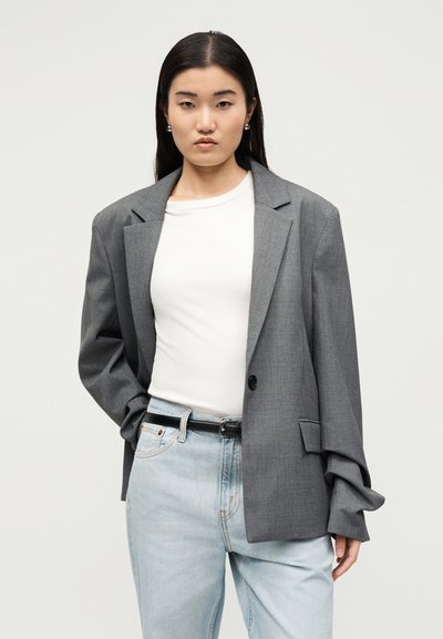 Γυναίκα με μακριά σκούρα μαλλιά, που φορά υπερμεγέθη γκρι σακάκι, λευκό πουκάμισο, ανοιχτό μπλε τζιν και μαύρη ζώνη, στέκεται μπροστά σε απλό φόντο.