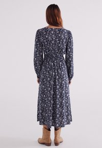 Marineblaues Blumendruck-Kleid mit langen Ärmeln, tailliertem Schnitt und knöchellangem Rock. Weicher Stoff und dezente Puffärmel. Getragen mit hellbraunen Knöchelstiefeln.