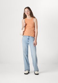 Oransje ribbet tanktop med sidelengs snøring, kombinert med lyseblå slengbukser og kraftige hvite sko, tilbehør med en liten veske.