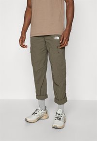 Olivengrønne cargo-bukser med glidelåslommer, oppbrettede cuffs og en hvit logo på livet, kombinert med grå sokker og lette sneakers.