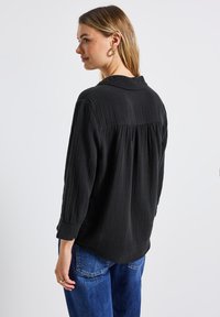 Svart långärmad skjorta med strukturerad tyg, knäppt krage, rynkad bakdetalj och uppvikta ärmar, kombinerad med blå högmidjade jeans.