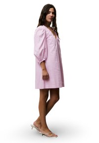 Rosenfarbenes Leinenkleid mit puffärmeligen und V-Ausschnitt, in mittlerer Oberschenkel-Länge. Getragen mit Sandalen mit Riemen und niedrigem Absatz.