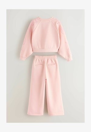 Világos rózsaszín pulcsi raglánujjakkal és nyomtatott díszítésekkel, párosítva a hozzá illő, széles lábú nadrággal, amely rugalmas derékpánttal és hátsó zsebekkel rendelkezik.