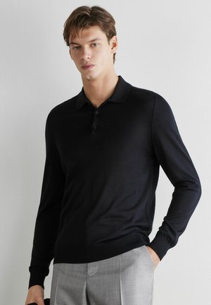 Männliches Model trägt ein schwarzes Langarm-Poloshirt und hellgraue Hose, steht vor einer schlichten weißen Wand.