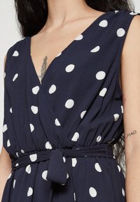 Ναυτικό φόρεμα με λευκές βούλες, διαθέτει βαθύ V-λαιμόκοψη, κοντά μανίκια, τονισμένη μέση και λεπτομέρεια με κορδόνι. Μαλακή υφή.