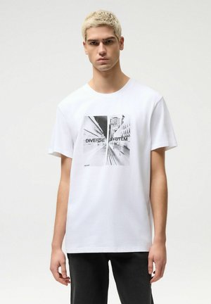 Młody mężczyzna z krótkimi blond włosami, ubrany w białą koszulkę z czarno-białym wizerunkiem miasta i napisem "DIVERSE SYSTEM", stoi na jednolitym tle.