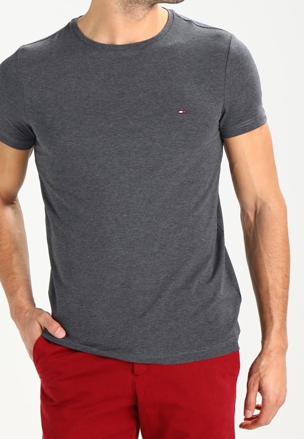 Hombre vistiendo una camiseta de manga corta gris oscuro lisa con un pequeño logo en el pecho y pantalones rojos, de pie con una mano en el bolsillo.