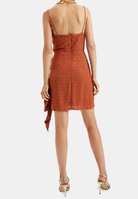 Robe sans manches couleur rouille avec un corsage ajusté et un ourlet fluide, présentant un motif texturé et un détail noué à la taille.