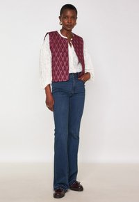 Donna che indossa una blusa bianca testurizzata, un gilet di raso bordeaux senza maniche, jeans a vita alta e svasati blu, e scarpe marroni scure, in piedi davanti a uno sfondo semplice.