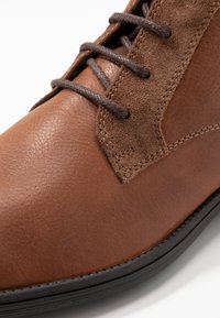 Brun läderstövel med mockadetaljer, rund tå, texturerad yta och kontrasterande mörka snören. Har svart gummisula och stickningsdetaljer.