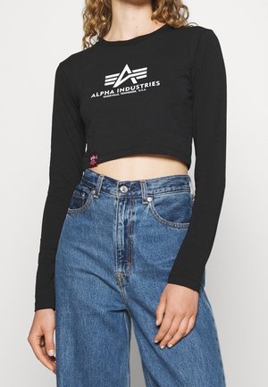 Osoba ubrana w czarny krótki top z długim rękawem z logo "Alpha Industries Knoxville Tennessee U.S.A." oraz niebieskie dżinsy z wysokim stanem.