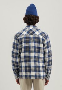 Geruite shirt in blauw, wit en grijze stoffen met een gestructureerde afwerking. Heeft een kraag en knoopsmanchetten, gedragen met een blauwe gebreide beanie.