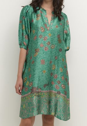 Vrouw draagt een groene jurk tot op de knie met pofmouwen, een bloemen- en patroonprint, en een V-hals.