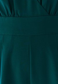 Teal jumpsuit med fitted talje, plisseret kjole og glat, struktureret stof. Syninger langs sømmene tilføjer definition.