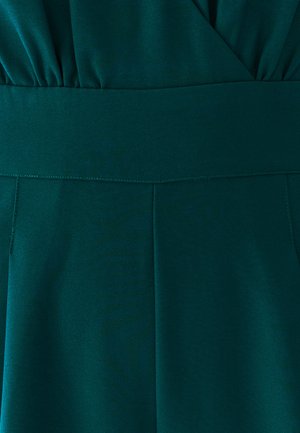 Niebieski kombinezon z dopasowanym wcięciem w talii, plisowanym przodem i gładkim, teksturowanym materiałem. Szczegóły przeszyć wzdłuż szwów dodają definicji.