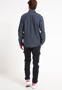 Chemise boutonnée bleu foncé à manches longues, associée à un pantalon cargo noir et des baskets blanches avec des accents rouges. Le tissu semble doux et texturé.