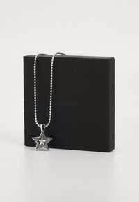 Sudraba zvaigzne ar teksturētu dizainu un pērļotu ķēdīti, novietota uz melnas kastes.