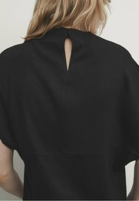 Schwarzes Kleidungsstück mit strukturierter Oberfläche, das eine Schlüssel-ausschnitt-Rückenpartie und einen lockeren, entspannten Schnitt aufweist. Schlichtes, modernes Design.