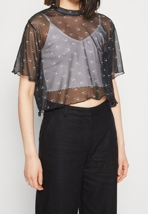 Kobieta w czarnym przezroczystym t-shircie Nike na szarym crop topie Nike i czarnych spodniach z wysokim stanem, stojąca na białym tle.