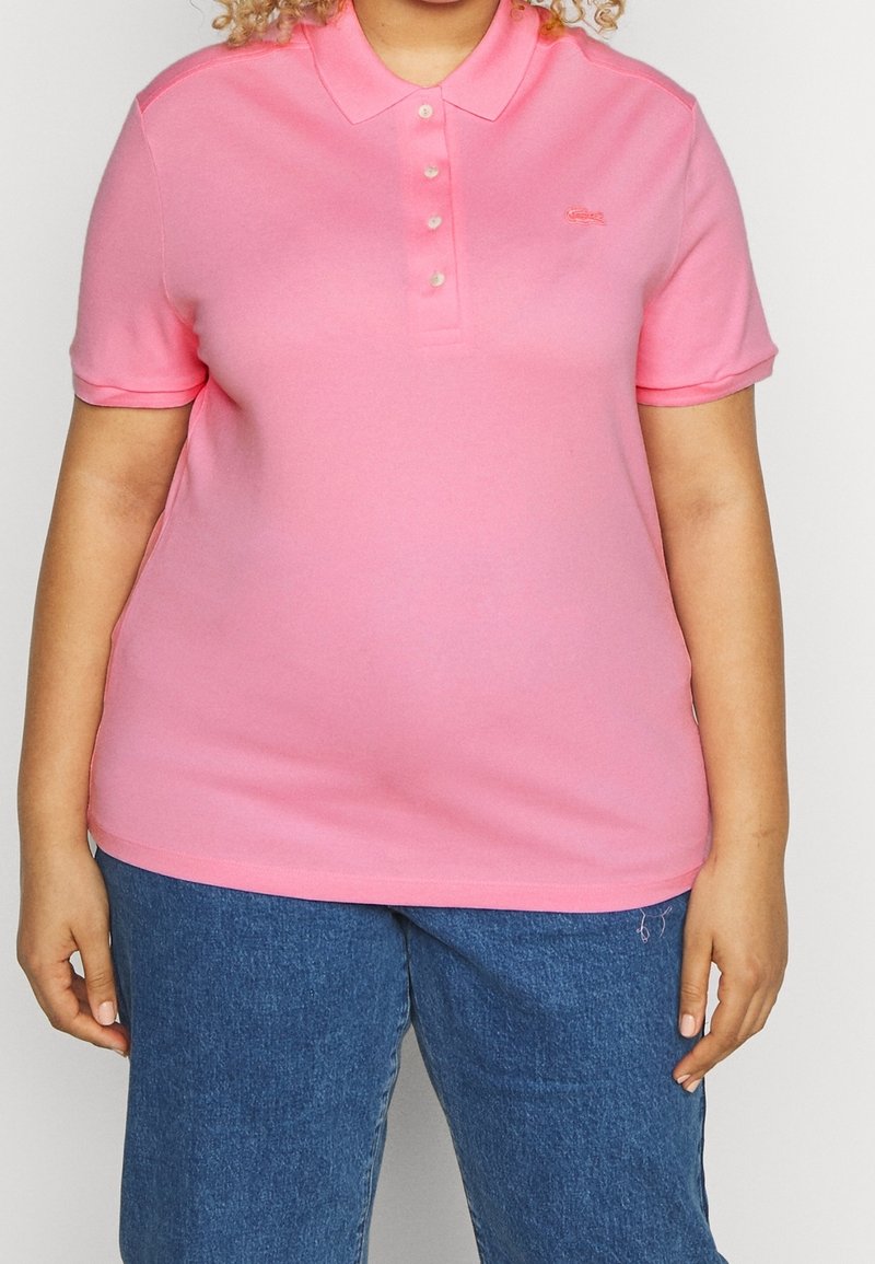 Personne portant un polo rose vif à manches courtes avec des boutons et un jean en denim bleu, debout devant un fond uni.