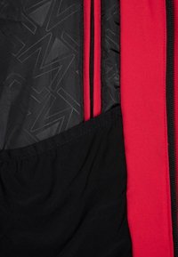 Acercamiento al interior de una chaqueta que muestra un bolsillo de malla negra, forro rojo, tela negra con diseño y un zipper negro vertical.