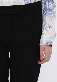 Sorte linbukser med en glat tekstur, der har en flad frontpanel, bælte loops og et lige ben design. Synlig blå blomsterbluse ærme.