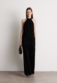 Μαύρο τοπ με halter σχέδιο και υφή, συνδυασμένο με φαρδιές, ρέουσες παντελόνες. Το μοντέλο κρατά μια μικρή μπορντό τσάντα. Απλό φόντο.