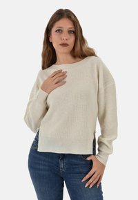 Maglione lavorato a maglia color bianco sporco con trama a coste, spalle scese e dettagli laterali di perle, abbinato a jeans blu.
