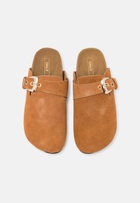 Sabots en daim marron sans lacets avec une large sangle, boucle réglable et semelle intérieure matelassée. Bout arrondi avec une texture lisse et mate.