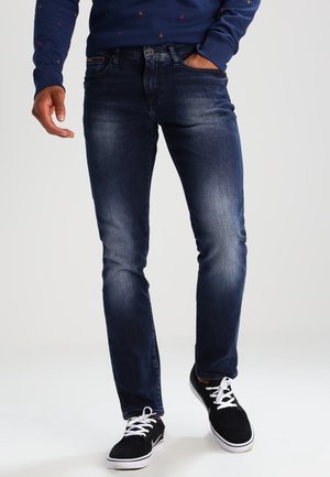 Mann trägt dunkelblaue Slim-Fit-Jeans mit leichter Waschung, schwarze Sneaker mit weißen Schnürsenkeln und ein dunkelblaues Langarmhemd mit kleinen Mustern.