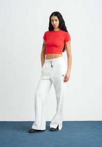 Sarkans īss T-krekls ar īsām piedurknēm un logotipu, kombinēts ar baltiem sporta biksēm, kurām ir aukla un šķēlumu atveres apakšmalā.