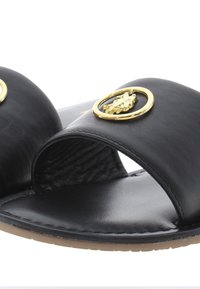 Černé kožené sandály na klínu mají široký pásek s zlatým kruhovým emblémem a texturovanou stélkou. Podešev je plochá s minimálním vzorkem.