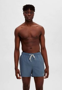 Homem de pele escura e dreadlocks curtos, sem camisa, usa calções de banho azuis com cordão, de pé contra um fundo claro e simples.