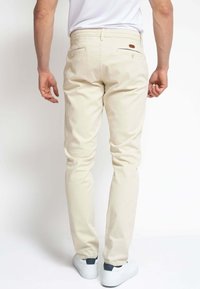 Chinos de algodón beige con un corte ajustado, que presentan bolsillos traseros y un pequeño parche de cuero marrón en la cinturilla, combinados con zapatillas blancas.