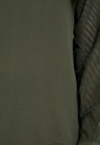 Vêtement vert olive avec une manche transparente et texturée présentant des rayures diagonales et de petits points en relief.