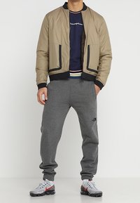 Tan nylon bomberjacka med svarta detaljer, tillsammans med grå sweatpants som har en logo. Sneakers har en grå och röd design.