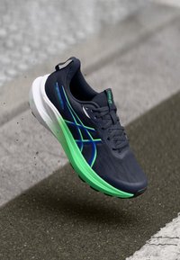 Σκούρο μπλε και πράσινο αθλητικό παπούτσι τρεξίματος με λευκή σόλα, να αιωρείται πάνω από υφασμάτινο πεζοδρόμιο, δείχνοντας λεπτομέρειες από το πλάι και την κορυφή.