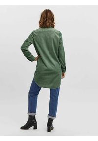 Camicia in corduroy verde, maniche lunghe, orlo curvo, abbinata a jeans con risvolto e stivaletti neri alla caviglia. Tessuto morbido, design casual.