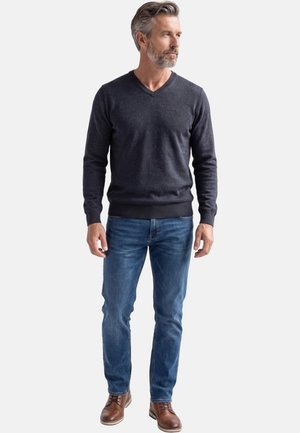 Homme aux cheveux gris et barbe portant un pull à col en V gris foncé, un jean bleu et des chaussures en cuir marron, debout et regardant vers la droite.