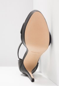 Chaussure noire à talon haut avec bout pointu, bride à la cheville et semelle beige, placée en angle contre un mur blanc.
