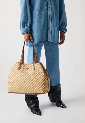 Personne portant une chemise en denim bleu et un jean, des bottes noires pointues, tenant un grand sac cabas tissé en paille avec des poignées en cuir marron.