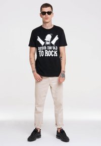 Black cotton t-shirt featuring a graphic print of Homer Simpson and text "Never Too Old To Rock." Worn with beige rolled-cuff pants and black sneakers.