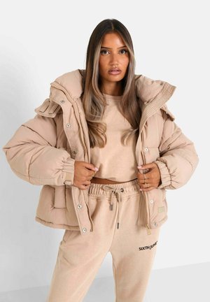Chaqueta acolchada beige con cuello alto, forro acanalado, puños elásticos y botones a presión. Usada sobre un top corto beige a juego y joggers.