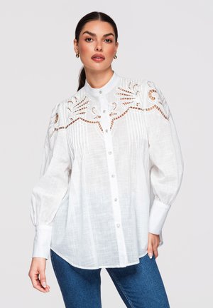 Kvinde iført en hvid skjorte med knapper og indviklet udskåret blonder på skuldre og øvre bryst, kombineret med blå jeans.