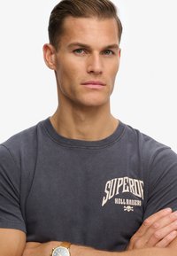 Camiseta de algodón gris oscuro con cuello redondo, que presenta las palabras "Superdry Hell Raisers" en beige claro, adornada con un gráfico de calavera.