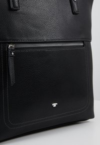 Sac cabas en cuir noir texturé avec poche zippée à l'avant, coutures visibles et petit logo de marque argenté sur le panneau avant inférieur.