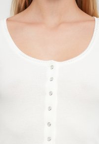 Κοντινή λήψη λευκού ριμπ πουκαμίσου με στρογγυλή λαιμόκοψη που διαθέτει κάθετη σειρά έξι ασημένιων κουμπιών τύπου σνάπ.